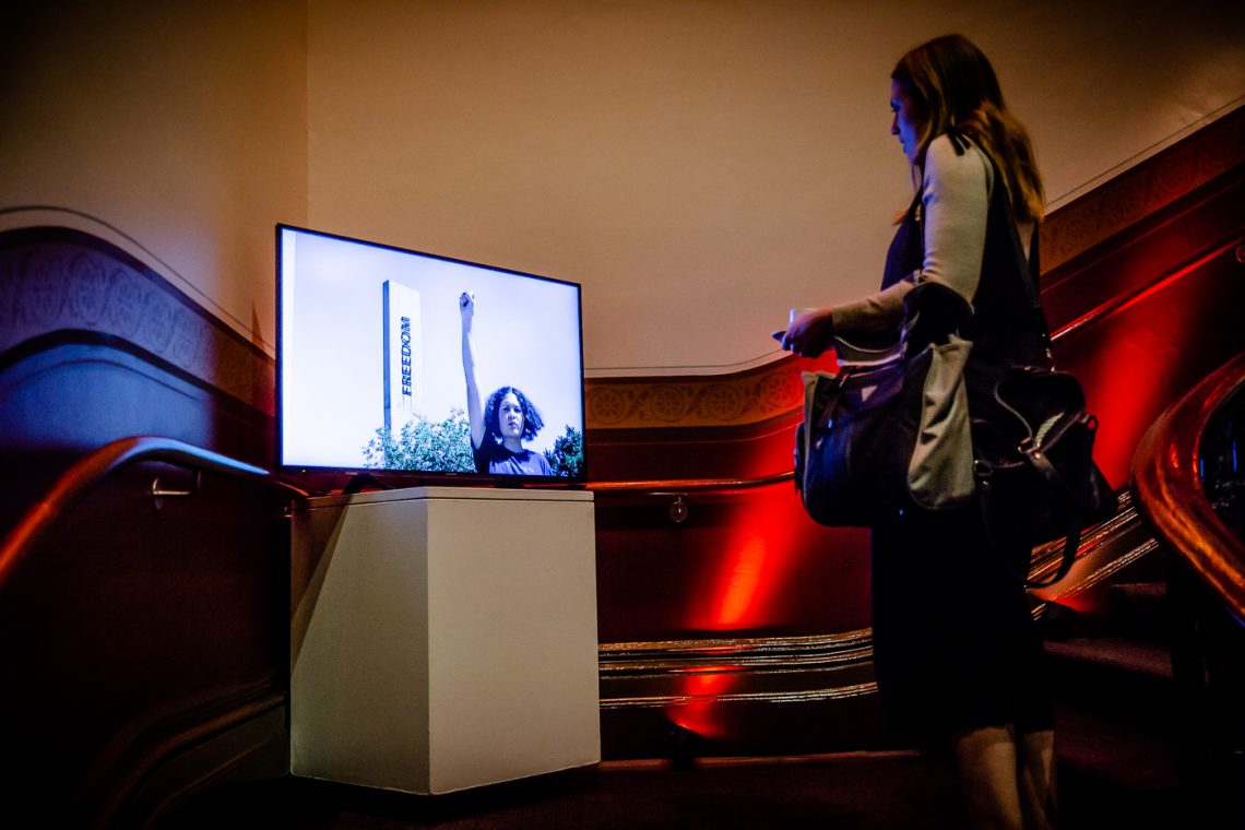 A woman is standing in a dramatically lit staircase watching a video on a flatscreen monitor of an artwork that features a woman with short curly hair, fist raised to the sky, standing next to a building that reads "freedom".